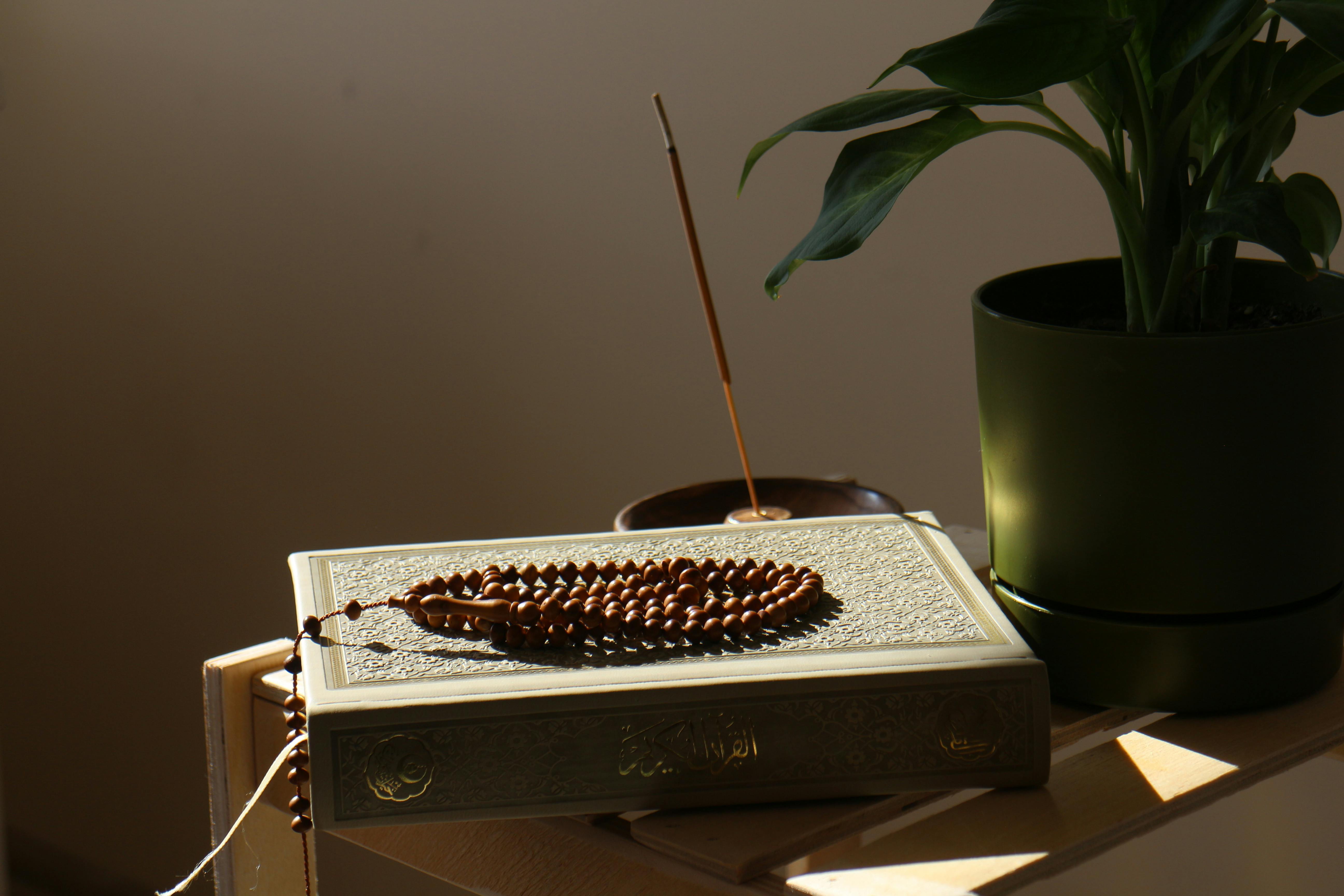 a Quran on top of a side table, with a rosary on top of the Quran. There is an incense stick, and a plant in a pot right next to it.