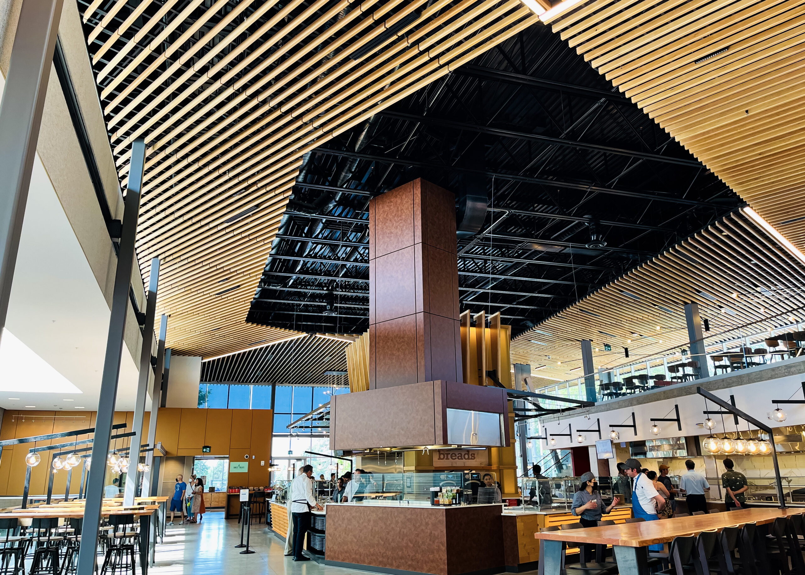 The SFU dining commons and food service workers interacting are pictured. The photo faces toward the ceiling, and there’s a sign that says “breads.”