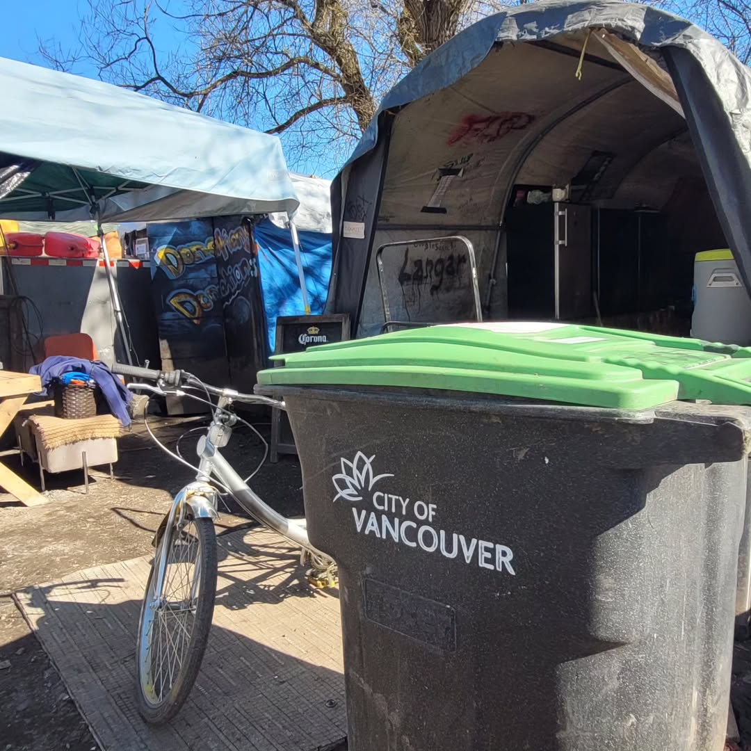 This is a photo of a shelter outside, with two large tents, a small fridge, a bench, and a bicycle featured. There is a garbage can that says “City of Vancouver” up close to the right side of the photo.