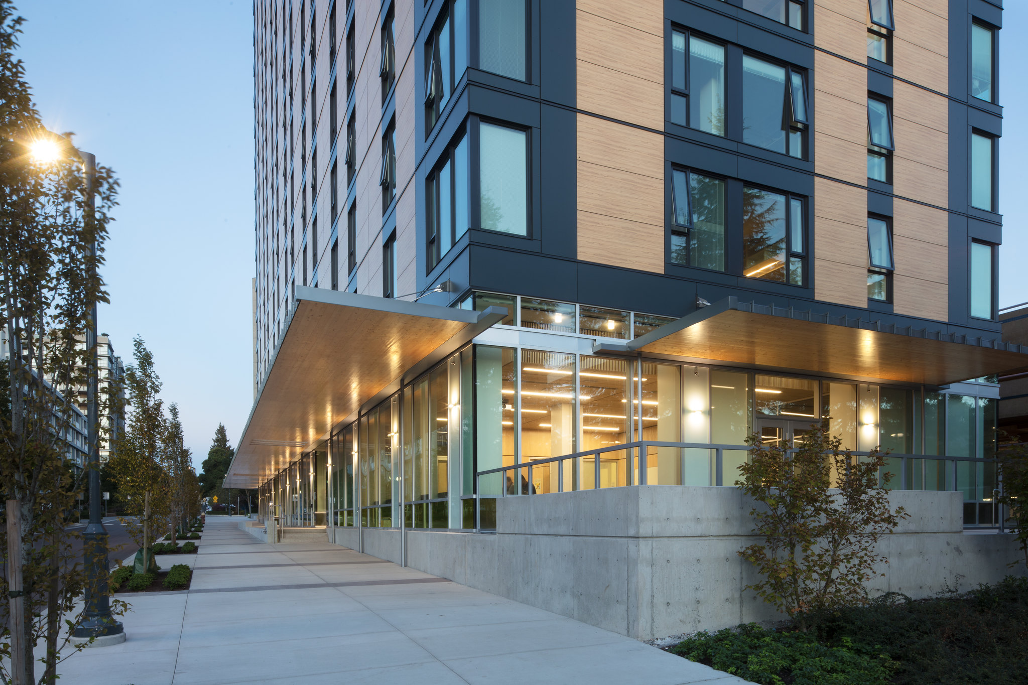 A UBC residence building is pictured from its corner. The building is black and brown, and has many large windows. The ground floor is lit up with warm orange lights, and a side street is pictured as well.