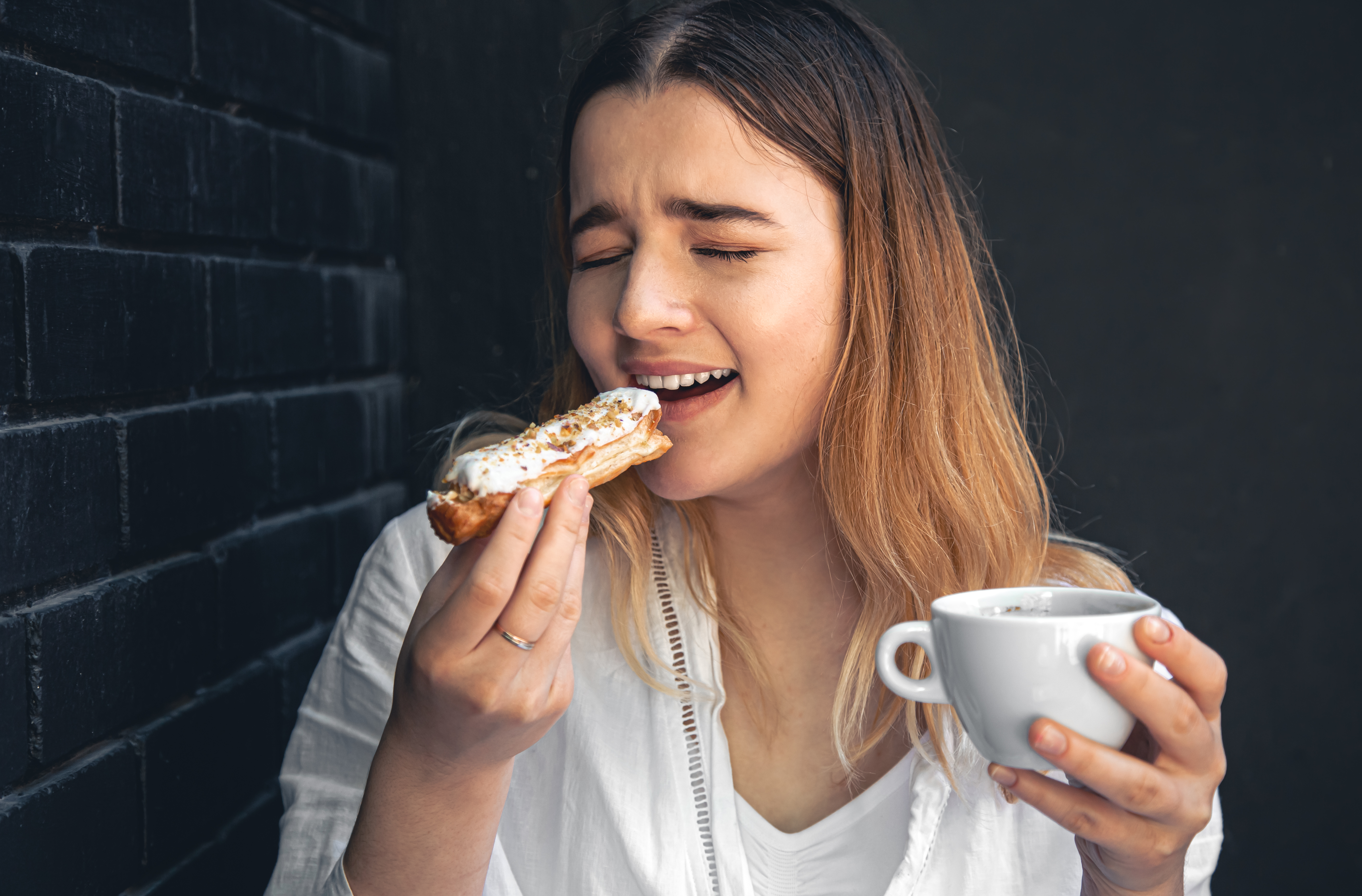 A woman holding a coffee cup and an éclair in her hands, enjoying her bites.