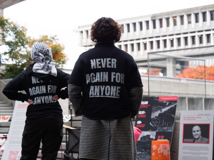 The back of two students’ shirts say, “never again for anyone.” One student is wearing a keffiyeh on their head, while a Palestinian flag and other posters are laid out on concrete steps in Main Mall.