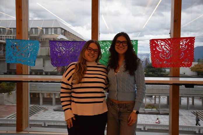 A photo of ALAS president Maria Clara Rezende (left) alongside Vice president Regina Zamira Sierra (right), smiling in front of the window from the SUB overlooking the Belzberg library. A string of papel picado (perforated tissue cut in intricate designs) hang along the width of the window behind them.