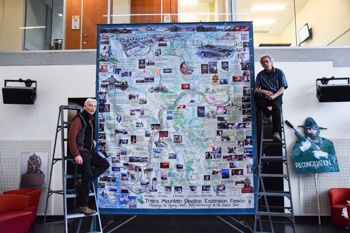 George Rammell and another man stand on ladders next to a tall piece of art. The art is a map with the title “The Trans Mountain Pipeline Expansion Fiasco: Mapping the Tipping Point, Fort McMurray to the Salish Sea” at the bottom. The map covers most of BC and features numerous photos, descriptions, and a snake as the Trans Mountain Pipeline.