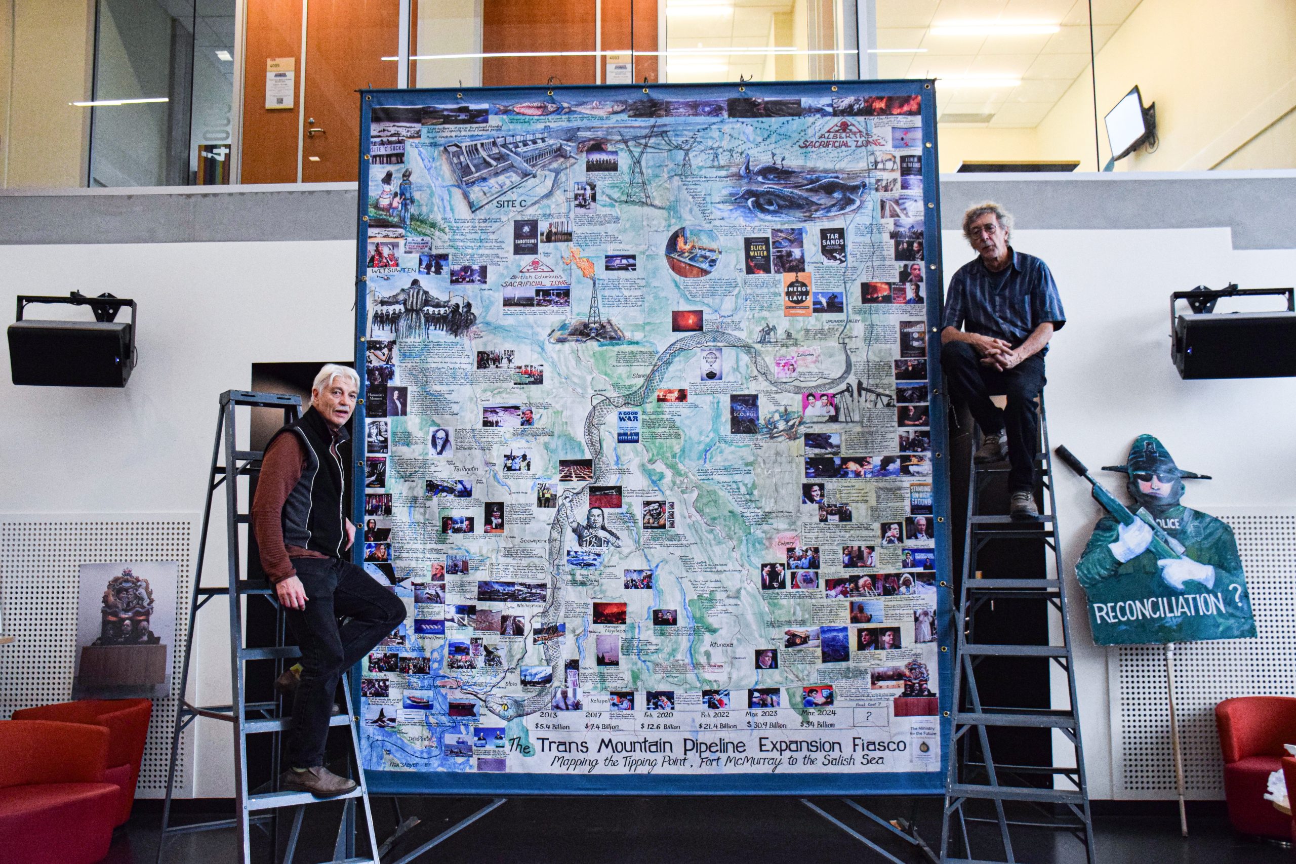 George Rammell and another man stand on ladders next to a tall piece of art. The art is a map with the title “The Trans Mountain Pipeline Expansion Fiasco: Mapping the Tipping Point, Fort McMurray to the Salish Sea” at the bottom. The map covers most of BC and features numerous photos, descriptions, and a snake as the Trans Mountain Pipeline.