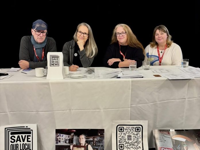 Four of Freshet’s founders sit at a long white table and smile for the camera. Their table has a small sign saying, “Save Our Local News.”