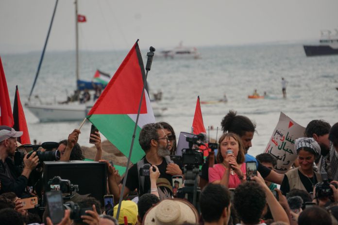 Greta Thunberg speaks among a close crowd of reporters and people holding up Palestinian flags. In the background, the ocean and a couple of boats are pictured, implying that this photo was taken sometime during the flotilla’s journey.
