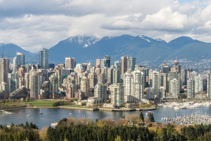 A wide shot of downtown Vancouver on a bright sunny day is pictured. False Creek and the mountains are also pictured.