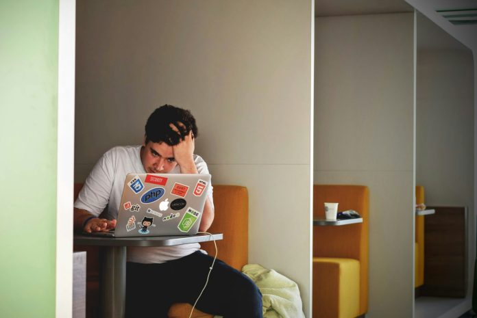 A student touches his head in frustration while looking at a computer at a desk.