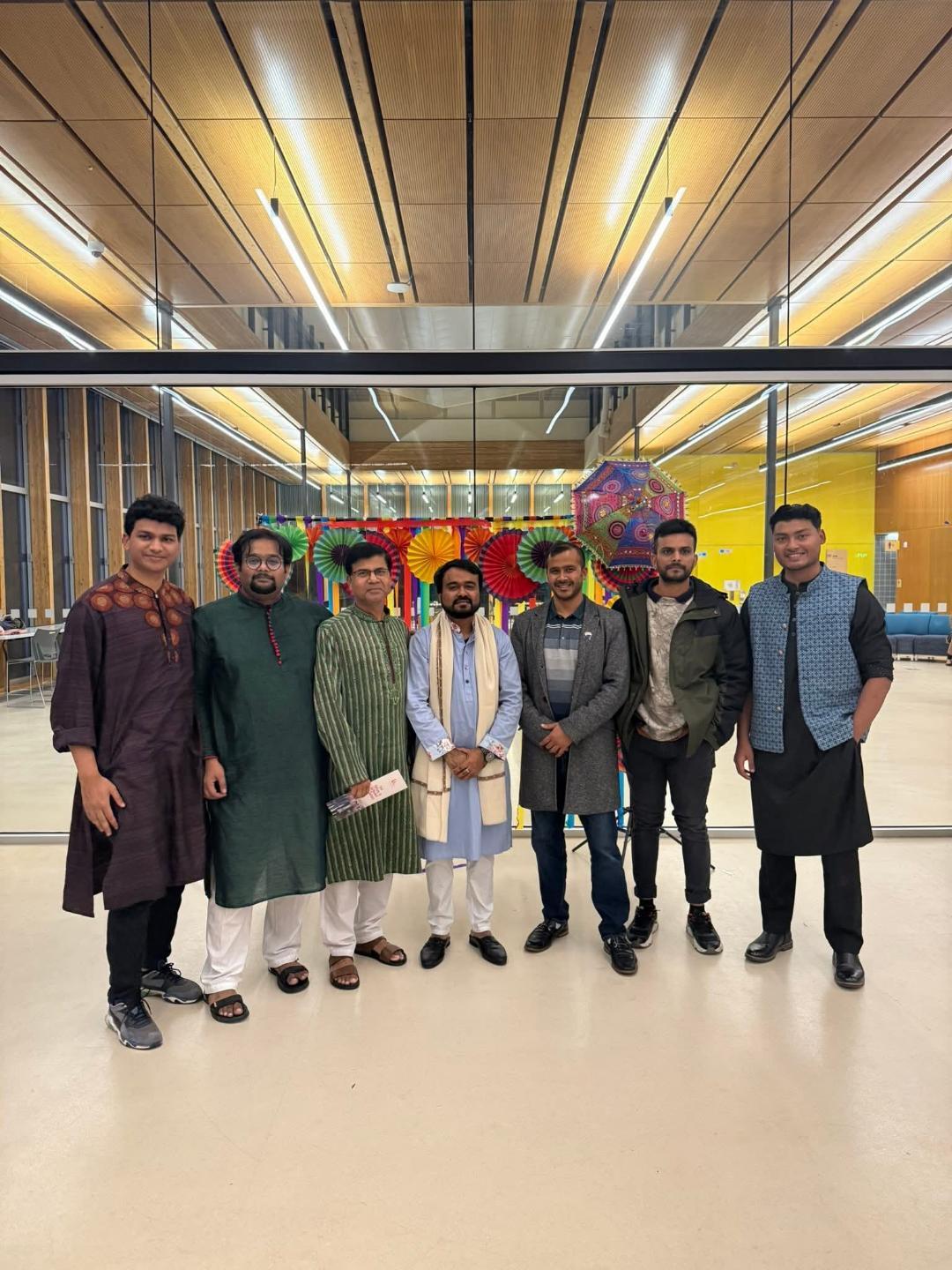 Seven men stand together and smile for the camera in traditional Bangladeshi attire in a brightly lit room with big windows.