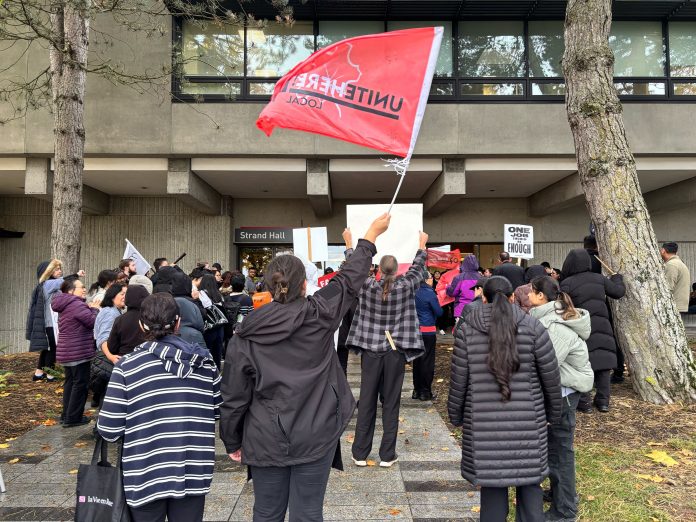 A crowd of people in jackets surrounds the entrance to a building, with the building’s sign being displayed near the entrance (“Strand Hall”). The person closest to the camera is waving a large red flag that says “UNITE HERE Local 40,” while others hold a few signs here and there as well. Another sign says, “ONE JOB SHOULD BE ENOUGH.”