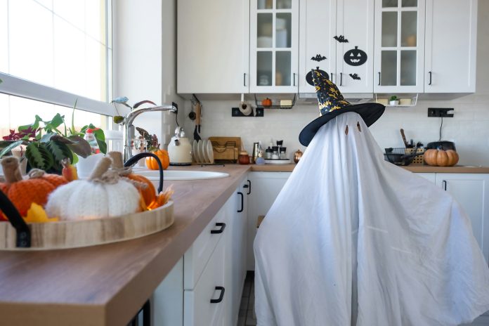 a kid dressed as a ghost, with a white sheet covering their body. They’re standing in a kitchen. With decorative pumpkins around the kitchen.