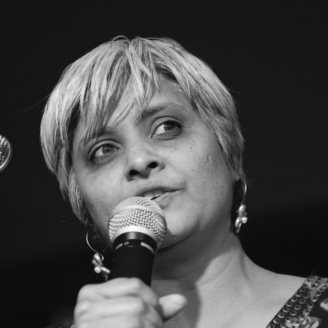 Pragna Patel is speaking into a microphone and looking off into the audience in this black and white headshot. She has short hair and hoop earrings.