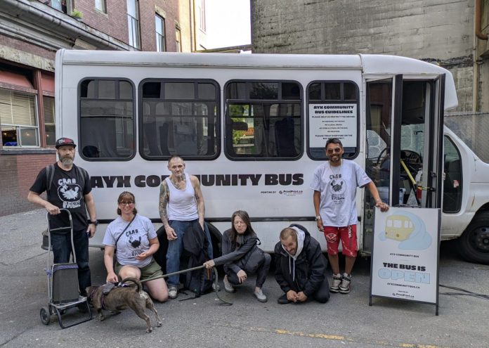 Six Ayx Bus workers and advocates stand outside the Ayx Bus with a sign that says, “the bus is open.” The white bus is about the size of a TransLink community shuttle.