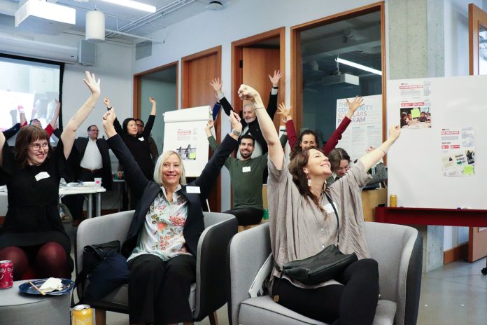 Ten people raise their arms in the air in celebration. Some of them sit in chairs, while others stand near the back of the room.