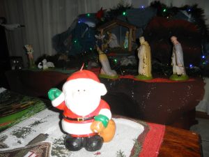A santa Claus action figure standing in front of a traditional nativity scene on display in someone’s house, where the three wisemen are lined up in front of a barn. The barn is decorated with garland and twinkling christmas lights. 