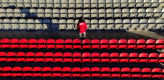 SFU moves forward to leave the NCAA A student wearing an SFU sweatshirt turns their back to the camera as they stand in the middle of the SFU Stadium stands. The photo is solemn and expresses loss, with the key colour being red for SFU’s Red Leafs.