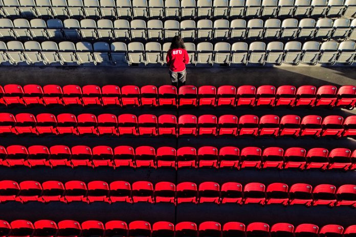 A student wearing an SFU sweatshirt turns their back to the camera as they stand in the middle of the SFU Stadium stands. The photo is solemn and expresses loss, with the key colour being red for SFU’s Red Leafs.
