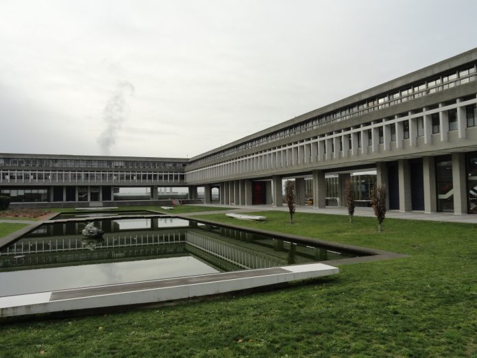 The Academic Quadtrangle at SFU is pictured on a cloudy day. The grey tone of the building, sky, and pond stands in stark contrast to the dark green grass.