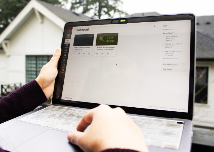 The Canvas dashboard is open on a student’s laptop. The student holds the side of the laptop’s screen with their left hand, while the other types.
