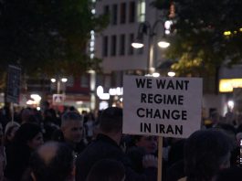 a crowd of protesters. One among them is holding a sign that reads: “We Want Regime Change in Iran.”