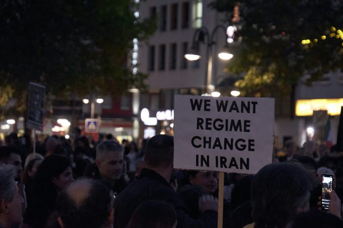 a crowd of protesters. One among them is holding a sign that reads: “We Want Regime Change in Iran.”