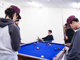 a photo of a group of people. One person is sitting down and is working on their tablet. While the others are congregated over a game of pool.