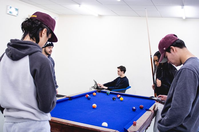 a photo of a group of people. One person is sitting down and is working on their tablet. While the others are congregated over a game of pool.