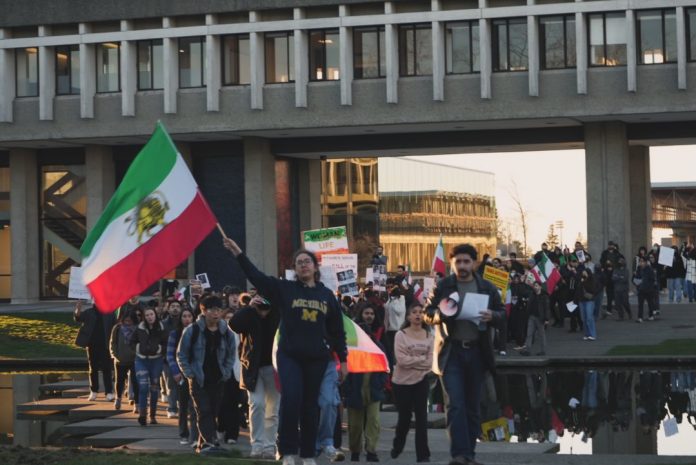 A large group of students march through the middle of the Academic Quadrangle with Iranian flags and signs at sunset.