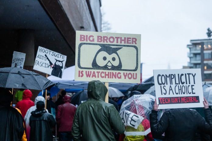 A crowd of people with their backs to the camera hold signs. Three of them read, “big brother is watching you,” “complicity is a choice: tech isn’t neutral,” and “Hootsuite aids fascism” with the Hootsuite owl logo.