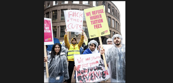 A group of protestors in ponchos smile for the camera and and hold their neon signs. Some of them read, “we refuse higher tuition fees,” “f#ck the cuts,” and “we are students, not your revenue.”