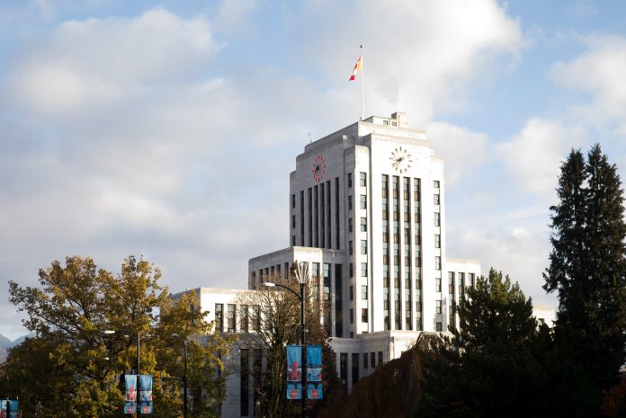 The Vancouver City Hall building is pictured on a bright, sunny day surrounded by trees and street lamps.