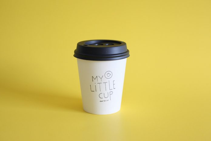 A photo of a to-go cup of coffee. The background is bright yellow. And the cup has “My Little Cup,” written on it.