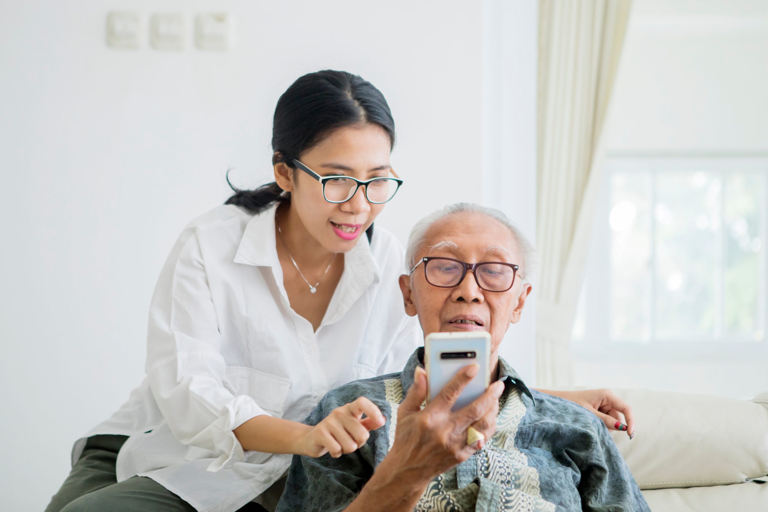 a woman helping her grandfather use his phone. The grandfather is holding the phone in his hand, while she is explaining how to navigate the phone.