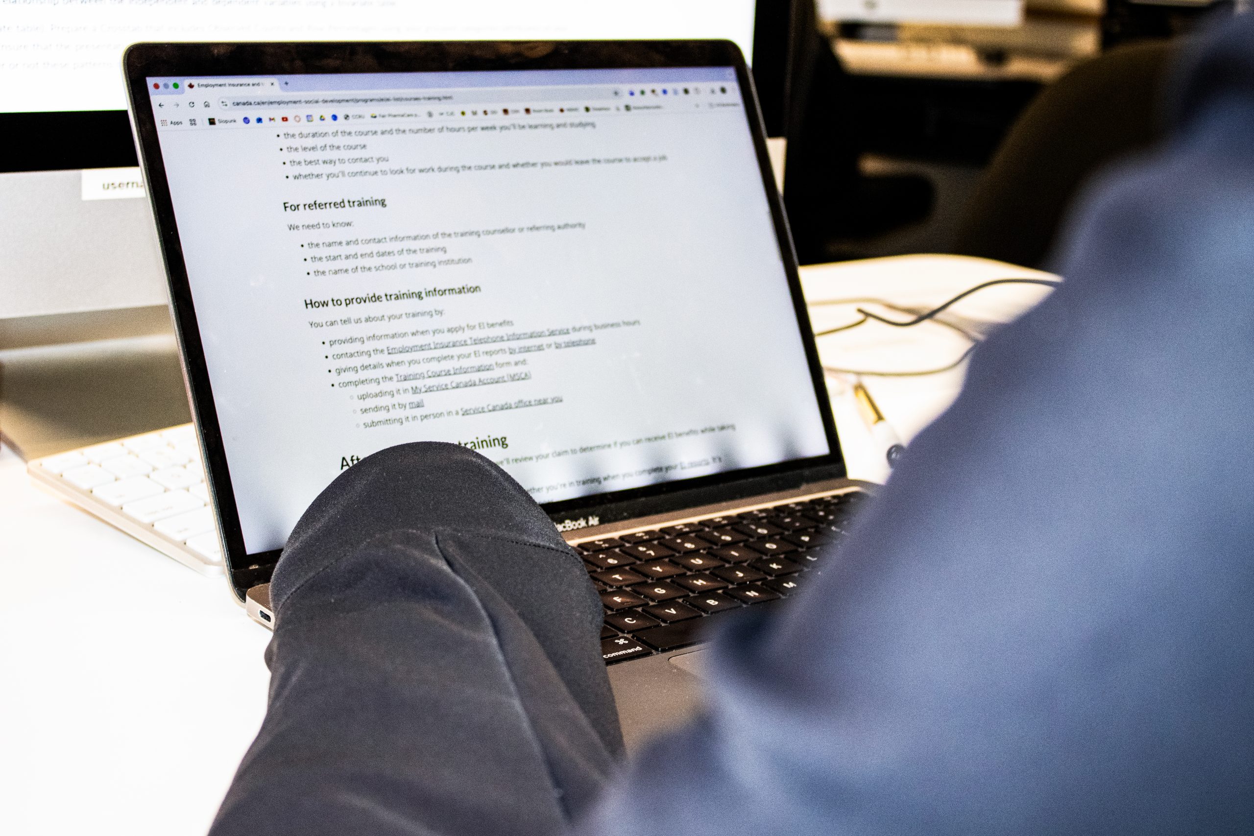 a person sitting in front of their laptop. The screen displays the website for employment insurance, on the training section of the website. One can only see the person’s arms in the photo.