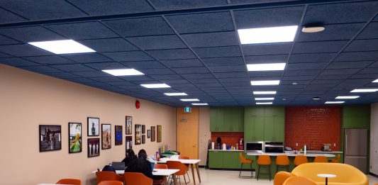 SFU debuts new Black Student Centre Bright hues of orange, blue, green, and yellow colour the chairs, ceiling, and cabinets of the Black Student Centre. Cushion-y seats are on the right, a green kitchen is in the back, and students sit in long desks near a row of photos on the left.
