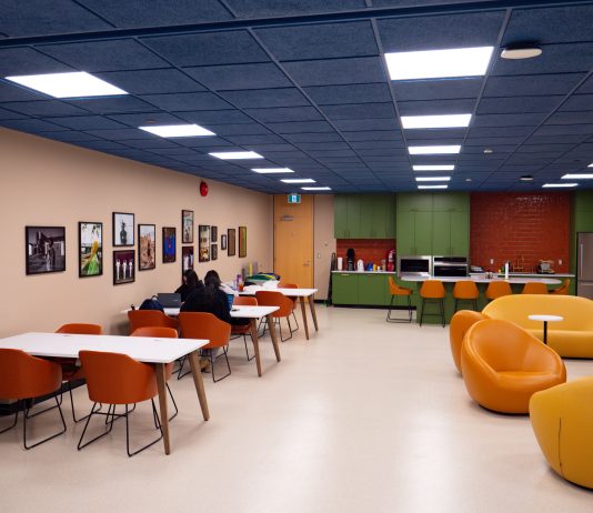 SFU debuts new Black Student Centre Bright hues of orange, blue, green, and yellow colour the chairs, ceiling, and cabinets of the Black Student Centre. Cushion-y seats are on the right, a green kitchen is in the back, and students sit in long desks near a row of photos on the left.