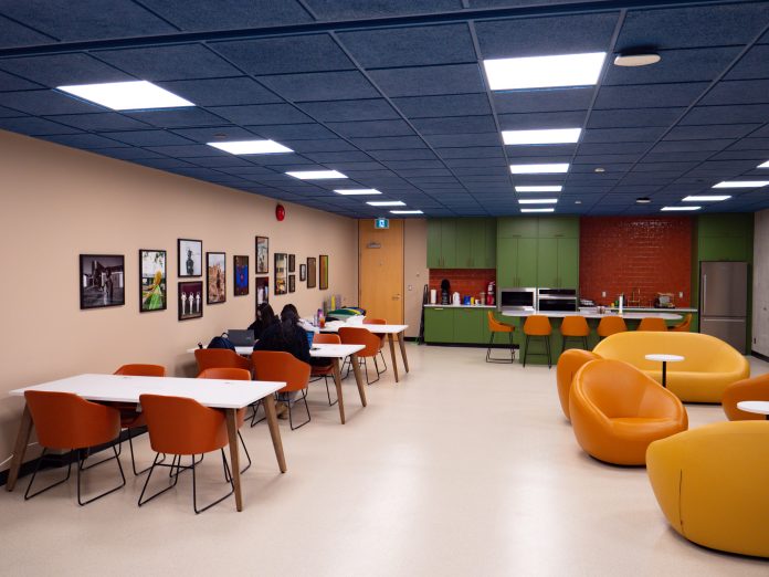 Bright hues of orange, blue, green, and yellow colour the chairs, ceiling, and cabinets of the Black Student Centre. Cushion-y seats are on the right, a green kitchen is in the back, and students sit in long desks near a row of photos on the left.