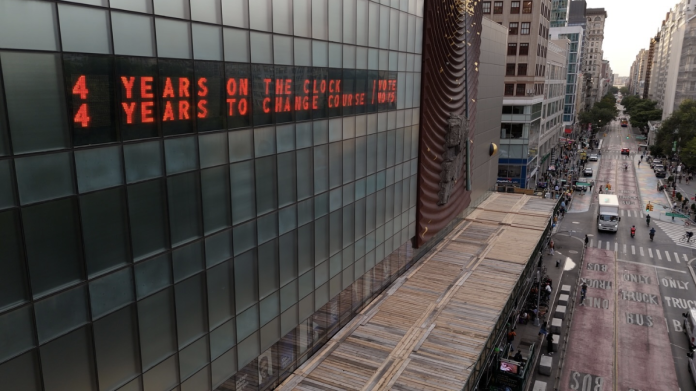 a photo of the art installation of the Climate Clock in New York City. The image shows the digital clock, positioned on a building, and parts of the street. The clock’s screen reads “4 years on the clock, 4 years to change course.”
