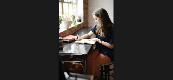A woman calmly journaling on a wooden desk in front of a sunny window.