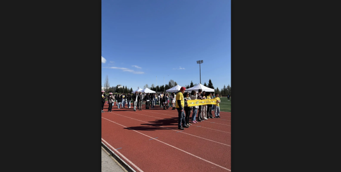Students, cancer survivors, and other participants stand together on the track of the Terry Fox Field. Some of them are wearing yellow shirts for the event.