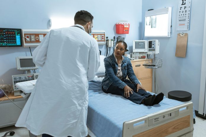 a woman during a doctor’s visit at the hospital. She sits on top of a hospital bed, and the doctor is standing to her right, asking her questions.