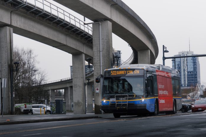 The 501 Surrey Ctrl bus drives along a road, below SkyTrain infrastructure.
