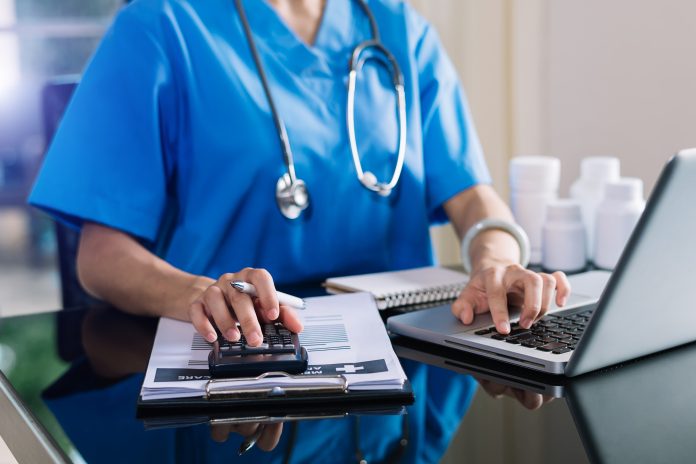 Stock image of a health-care worker in their scrubs, stethoscope visible. They are typing on a computer.