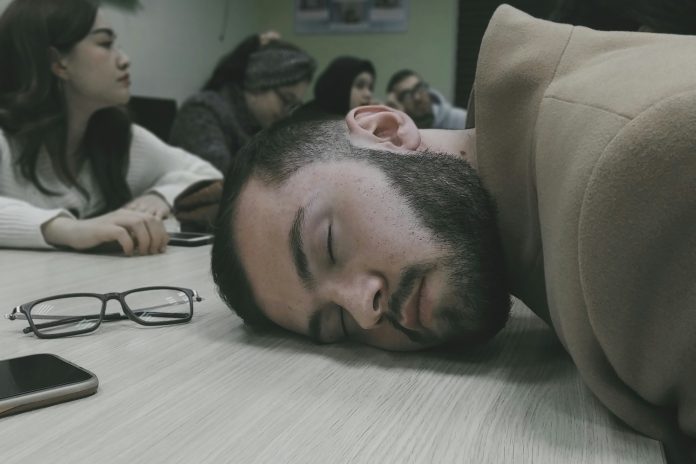A student resting his head on a table sleeping, black reading glasses sitting beside him