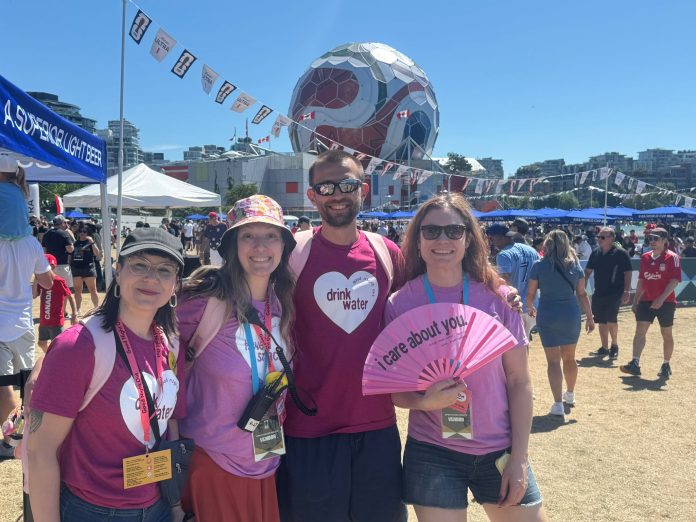 Four Good Night Out Vancouver volunteers pose together in front of FIFA soccer-ball-decorated Science World, dressed in pink t-shirts with white hearts embossed with various messages including “drink water.” Streamers and people are in the background, indicating a festival environment.