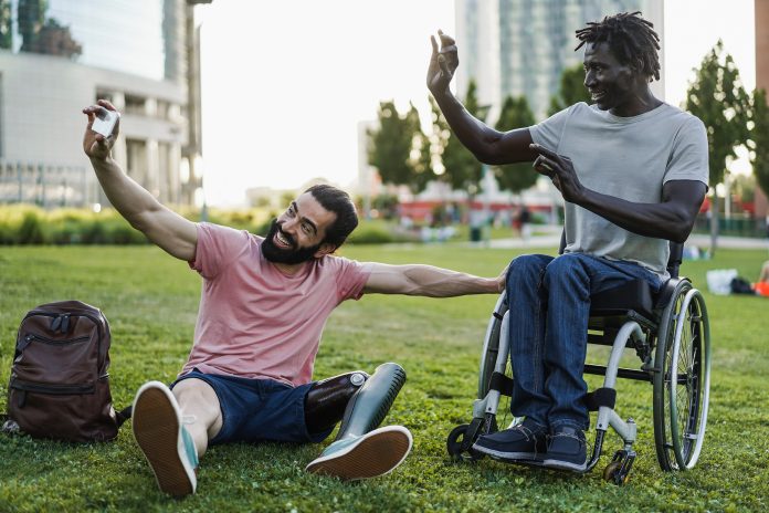 Two men, one sitting in a wheel chair and the other with an artificial leg, are taking a selfie outside on the grass