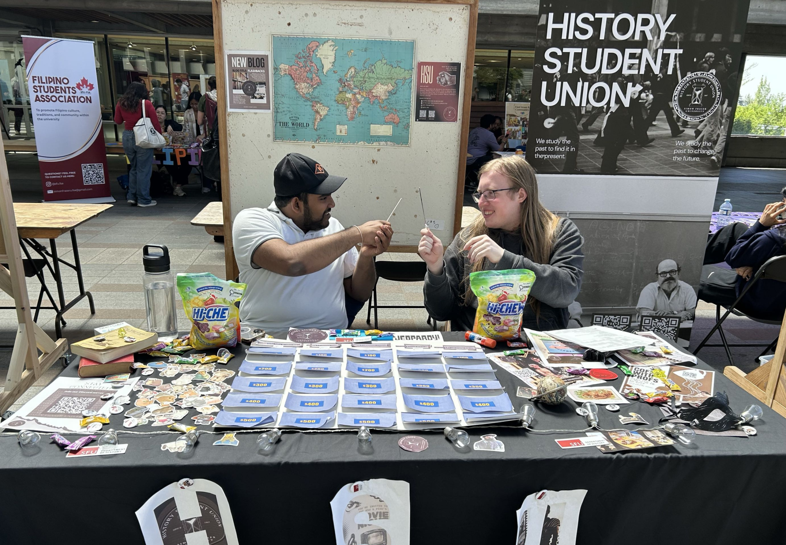 Two friends sit behind the booth table of the History Student Union. They interact playfully with each other.