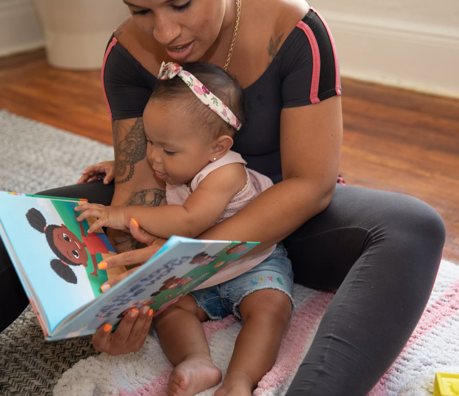 A parent reads to their infant the illustrated book Dahlia & Friends: All The Toys In The World. The open page shows a young Black girl.